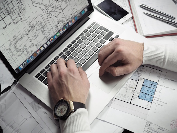 man working on architecture project on laptop on top of architectural paper plans